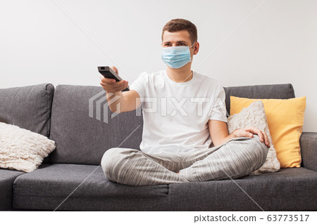 Young man in medical mask sits on couch and wathing television during quarantine. Young man in medical mask sits on couch and wathing television during quarantine. 63773517