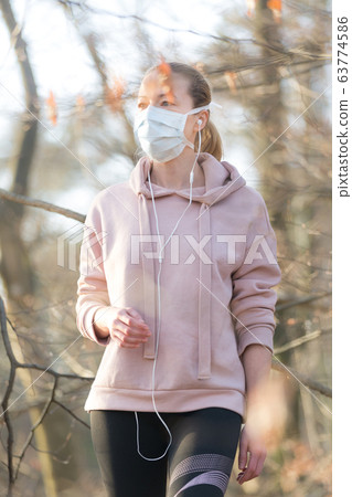 Portrait of caucasian sporty woman wearing medical protection face mask while walking in park, relaxing and listening to music. Corona virus, or Covid-19, is spreading all over the world 63774586