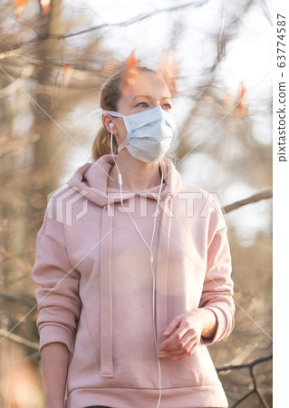 Portrait of caucasian sporty woman wearing medical protection face mask while walking in park, relaxing and listening to music. Corona virus, or Covid-19, is spreading all over the world Portrait of caucasian sporty woman wearing medical protection face mask while walking in park, relaxing and listening to music. Corona virus, or Covid-19, is spreading all over the world 63774587