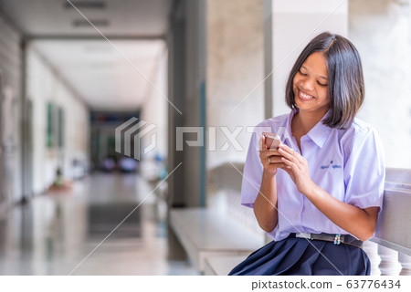Smiling Asian female high school student in white uniform is enjoying social media on her smartphone. Smiling Asian female high school student in white uniform is enjoying social media on her smartphone. 63776434
