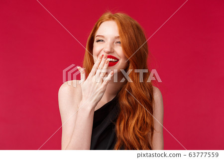 Close-up portrait of pretty elegant young european woman with long curly red hair, black dress and evening makeup, laughing over something funny looking away and cover smile with palm Close-up portrait of pretty elegant young european woman with long curly red hair, black dress and evening makeup, laughing over something funny looking away and cover smile with palm 63777559