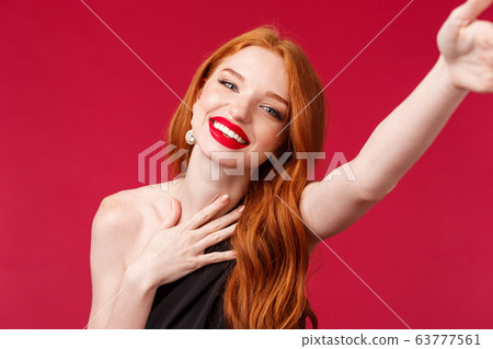 Close-up portrait of gorgeous smiling, happy redhead woman enjoying her birthday party, taking selfie for social media in stylish black dress and prom look with red lipstick, red background Close-up portrait of gorgeous smiling, happy redhead woman enjoying her birthday party, taking selfie for social media in stylish black dress and prom look with red lipstick, red background 63777561