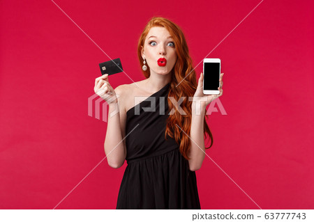 Fascinated and excited good-looking european woman with long curly ginger hair, wear black dress, holding mobile phone, showing smartphone display and credit card, shopping and internet concept 63777743