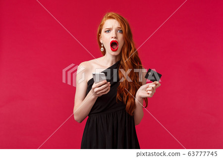 Portrait of complaining, disappointed gloomy clingy girlfriend with ginger long hair, feel unfair and jealous, holding credit card and smartphone, wanting buy something in internet, red background 63777745