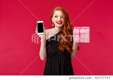 Portrait of excited and amused smiling, wondered redhead woman didnt expact receive gift so fast, showing mobile phone application, online delivery app, hold pink present over red background 63777757