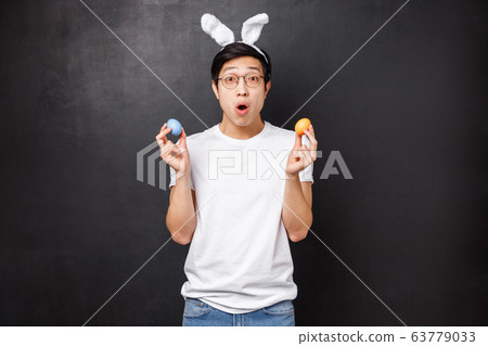 Holidays, party and Easter concept. Amazed young asian guy in rabbit ears holding painted eggs as learning girlfriend traditions, open mouth fascinated get to know new culture, black background 63779033