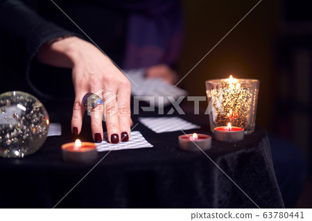 Close-up of female fortuneteller's hand with fortune-telling ring on cards at table with candles in dark room, blurred background Close-up of female fortuneteller's hand with fortune-telling ring on cards at table with candles in dark room, blurred background 63780441