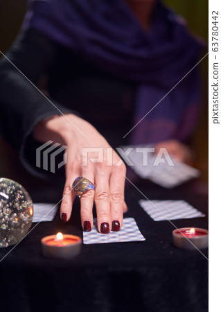 Close-up of woman fortuneteller's hand with fortune-telling cards at table with candles in dark room Close-up of woman fortuneteller's hand with fortune-telling cards at table with candles in dark room 63780442