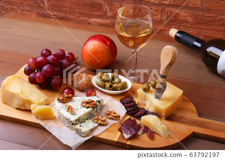 Assortment of cheese with fruits, grapes, nuts, glass with wine and cheese knife on a wooden serving tray. Assortment of cheese with fruits, grapes, nuts, glass with wine and cheese knife on a wooden serving tray. 63792197