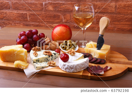 Assortment of cheese with fruits, grapes, nuts, glass with wine and cheese knife on a wooden serving tray. Assortment of cheese with fruits, grapes, nuts, glass with wine and cheese knife on a wooden serving tray. 63792198