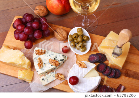 Assortment of cheese with fruits, grapes, nuts, glass with wine and cheese knife on a wooden serving tray. Assortment of cheese with fruits, grapes, nuts, glass with wine and cheese knife on a wooden serving tray. 63792200