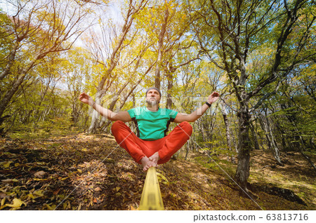 An adult bearded barefoot man sits and meditates on a slackline in the autumn forest. Yoga and balance exercises. Spiritual health An adult bearded barefoot man sits and meditates on a slackline in the autumn forest. Yoga and balance exercises. Spiritual health 63813716