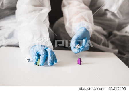 Two people in protective white translucent suit, blue rubber gloves, medical mask sit on sofa at home and play board games, roll dice. Stay home during coronavirus pandemic covid. Two people in protective white translucent suit, blue rubber gloves, medical mask sit on sofa at home and play board games, roll dice. Stay home during coronavirus pandemic covid. 63819493