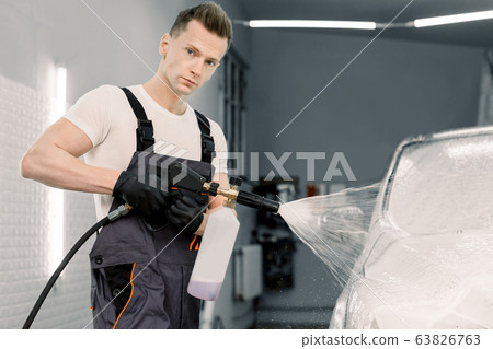 Close up portrait of car wash worker, hansome young man in protective overalls, cleaning automobile with high pressure water jet at car wash, spraying the cleaning foam 63826763