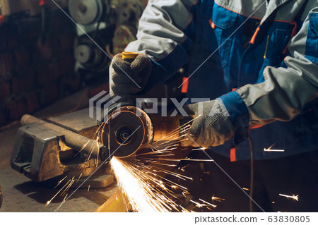 Locksmith in special clothes and goggles works in production. Metal processing with angle grinder. Sparks in metalworking 63830805