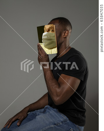 Be safe and read to become someone else - man covering face with book in face mask while reading on grey background Be safe and read to become someone else - man covering face with book in face mask while reading on grey background 63847035
