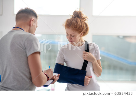 Male physiotherapist writing instructions, female patient standing in front of him in arm sling 63854433