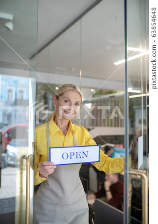 Blonde woman standing in doorway, holding sign saying open 63854648