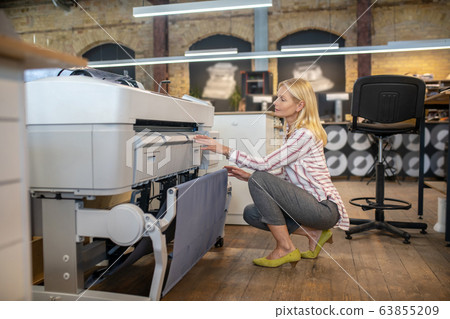 Blonde woman crouching at printing machine, checking something 63855209