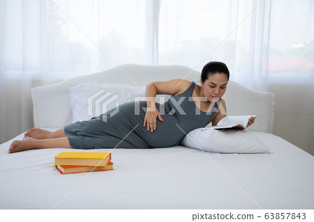 Asian Pregnant mother lying on bed and reading book Asian Pregnant mother lying on bed and reading book 63857843