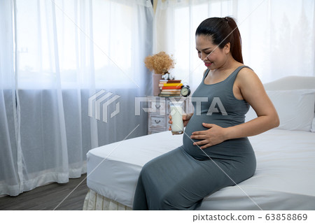 Asian Pregnant mother sitting on bed and holding milk glass for drinking Asian Pregnant mother sitting on bed and holding milk glass for drinking 63858869