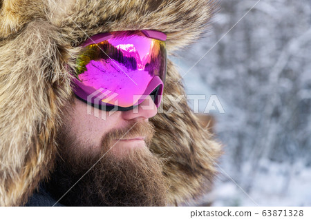 Close-up portrait of a bearded happy snowboarder skier in a ski mask with goggles and a fur big old-school hat on a background of a winter snowy mountains Close-up portrait of a bearded happy snowboarder skier in a ski mask with goggles and a fur big old-school hat on a background of a winter snowy mountains 63871328