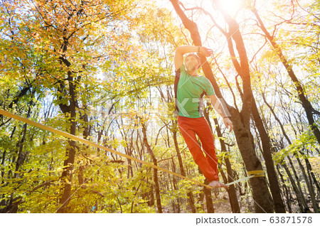 Wide angle male tightrope walker balancing barefoot on slackline in autumn forest. The concept of outdoor sports and active life of people aged Wide angle male tightrope walker balancing barefoot on slackline in autumn forest. The concept of outdoor sports and active life of people aged 63871578