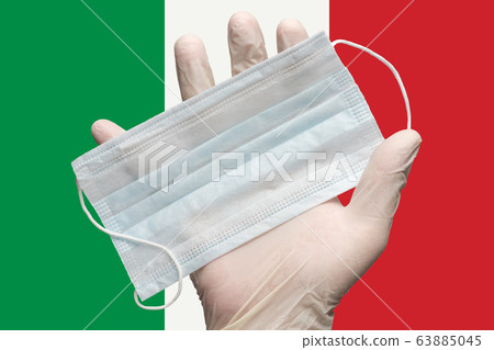 Doctor holds medical face mask in hand in white gloves on background colors flag of Italy. Concept pandemic insurance coronavirus Doctor holds medical face mask in hand in white gloves on background colors flag of Italy. Concept pandemic insurance coronavirus 63885045