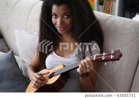 Dark-skinned girl sitting on sofa playing ukulele Dark-skinned girl sitting on sofa playing ukulele 63885751