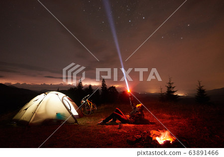 Woman resting at night camping near campfire, tourist tent, bicycle under evening sky full of stars 63891466