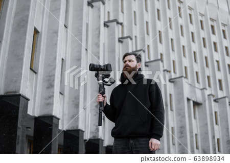 Bearded Professional videographer in black hoodie holding professional camera on 3-axis gimbal stabilizer. Filmmaker making a great video with a professional cinema camera. Cinematographer. Bearded Professional videographer in black hoodie holding professional camera on 3-axis gimbal stabilizer. Filmmaker making a great video with a professional cinema camera. Cinematographer. 63898934