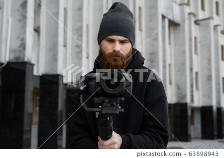 Bearded Professional videographer in black hoodie holding professional camera on 3-axis gimbal stabilizer. Filmmaker making a great video with a professional cinema camera. Cinematographer. 63898943