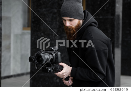 Bearded Professional videographer in black hoodie holding professional camera on 3-axis gimbal stabilizer. Filmmaker making a great video with a professional cinema camera. Cinematographer. Bearded Professional videographer in black hoodie holding professional camera on 3-axis gimbal stabilizer. Filmmaker making a great video with a professional cinema camera. Cinematographer. 63898949