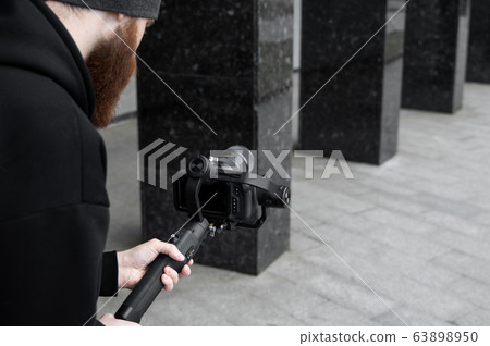 Bearded Professional videographer in black hoodie holding professional camera on 3-axis gimbal stabilizer. Filmmaker making a great video with a professional cinema camera. Cinematographer. Bearded Professional videographer in black hoodie holding professional camera on 3-axis gimbal stabilizer. Filmmaker making a great video with a professional cinema camera. Cinematographer. 63898950
