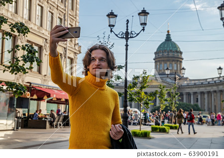 The handsome men does selfie, Look from outside, he dressed in a yellow sweater, a black raincoat or jacket is his hands, Bolshaya Konyushennaya street and Kazan Cathedral on background, sunny day The handsome men does selfie, Look from outside, he dressed in a yellow sweater, a black raincoat or jacket is his hands, Bolshaya Konyushennaya street and Kazan Cathedral on background, sunny day 63900191