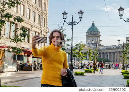 The handsome men does selfie, Look from outside, he dressed in a yellow sweater, a black raincoat or jacket is his hands, Bolshaya Konyushennaya street and Kazan Cathedral on background, sunny day The handsome men does selfie, Look from outside, he dressed in a yellow sweater, a black raincoat or jacket is his hands, Bolshaya Konyushennaya street and Kazan Cathedral on background, sunny day 63900202