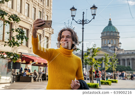 The handsome men does selfie, Look from outside, he dressed in a yellow sweater, a black raincoat or jacket is his hands, Bolshaya Konyushennaya street and Kazan Cathedral on background, sunny day The handsome men does selfie, Look from outside, he dressed in a yellow sweater, a black raincoat or jacket is his hands, Bolshaya Konyushennaya street and Kazan Cathedral on background, sunny day 63900216
