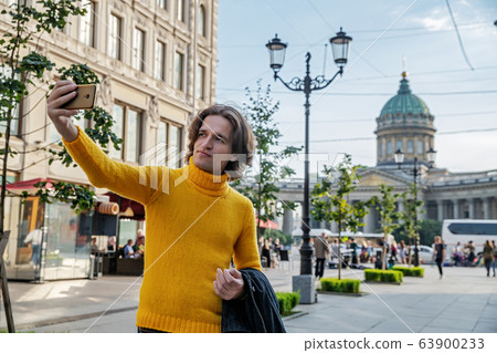 The handsome men does selfie, Look from outside, he dressed in a yellow sweater, a black raincoat or jacket is his hands, Bolshaya Konyushennaya street and Kazan Cathedral on background, sunny day The handsome men does selfie, Look from outside, he dressed in a yellow sweater, a black raincoat or jacket is his hands, Bolshaya Konyushennaya street and Kazan Cathedral on background, sunny day 63900233