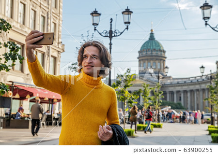The handsome men does selfie, Look from outside, he dressed in a yellow sweater, a black raincoat or jacket is his hands, Bolshaya Konyushennaya street and Kazan Cathedral on background, sunny day The handsome men does selfie, Look from outside, he dressed in a yellow sweater, a black raincoat or jacket is his hands, Bolshaya Konyushennaya street and Kazan Cathedral on background, sunny day 63900255