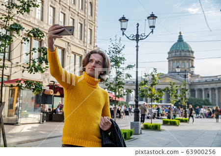 The handsome men does selfie, Look from outside, he dressed in a yellow sweater, a black raincoat or jacket is his hands, Bolshaya Konyushennaya street and Kazan Cathedral on background, sunny day The handsome men does selfie, Look from outside, he dressed in a yellow sweater, a black raincoat or jacket is his hands, Bolshaya Konyushennaya street and Kazan Cathedral on background, sunny day 63900256