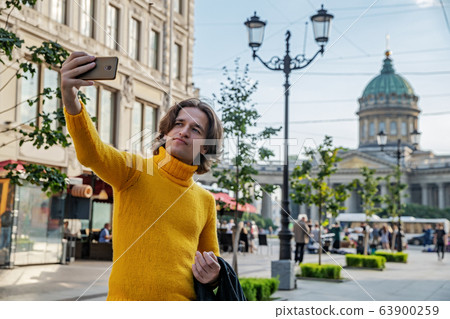 The handsome men does selfie, Look from outside, he dressed in a yellow sweater, a black raincoat or jacket is his hands, Bolshaya Konyushennaya street and Kazan Cathedral on background, sunny day The handsome men does selfie, Look from outside, he dressed in a yellow sweater, a black raincoat or jacket is his hands, Bolshaya Konyushennaya street and Kazan Cathedral on background, sunny day 63900259