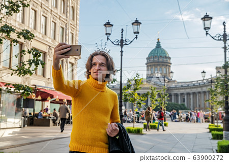 The handsome men does selfie, Look from outside, he dressed in a yellow sweater, a black raincoat or jacket is his hands, Bolshaya Konyushennaya street and Kazan Cathedral on background, sunny day The handsome men does selfie, Look from outside, he dressed in a yellow sweater, a black raincoat or jacket is his hands, Bolshaya Konyushennaya street and Kazan Cathedral on background, sunny day 63900272