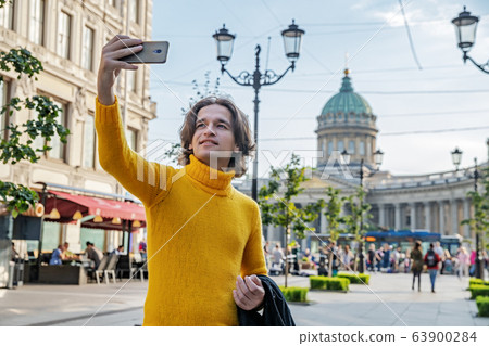 The handsome men does selfie, Look from outside, he dressed in a yellow sweater, a black raincoat or jacket is his hands, Bolshaya Konyushennaya street and Kazan Cathedral on background, sunny day The handsome men does selfie, Look from outside, he dressed in a yellow sweater, a black raincoat or jacket is his hands, Bolshaya Konyushennaya street and Kazan Cathedral on background, sunny day 63900284