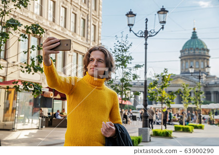 The handsome men does selfie, Look from outside, he dressed in a yellow sweater, a black raincoat or jacket is his hands, Bolshaya Konyushennaya street and Kazan Cathedral on background, sunny day The handsome men does selfie, Look from outside, he dressed in a yellow sweater, a black raincoat or jacket is his hands, Bolshaya Konyushennaya street and Kazan Cathedral on background, sunny day 63900290
