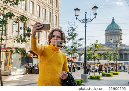 The handsome men does selfie, Look from outside, he dressed in a yellow sweater, a black raincoat or jacket is his hands, Bolshaya Konyushennaya street and Kazan Cathedral on background, sunny day The handsome men does selfie, Look from outside, he dressed in a yellow sweater, a black raincoat or jacket is his hands, Bolshaya Konyushennaya street and Kazan Cathedral on background, sunny day 63900293