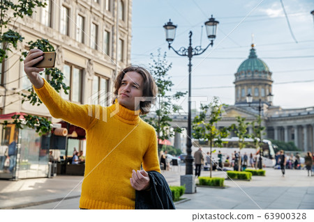 The handsome men does selfie, Look from outside, he dressed in a yellow sweater, a black raincoat or jacket is his hands, Bolshaya Konyushennaya street and Kazan Cathedral on background, sunny day The handsome men does selfie, Look from outside, he dressed in a yellow sweater, a black raincoat or jacket is his hands, Bolshaya Konyushennaya street and Kazan Cathedral on background, sunny day 63900328