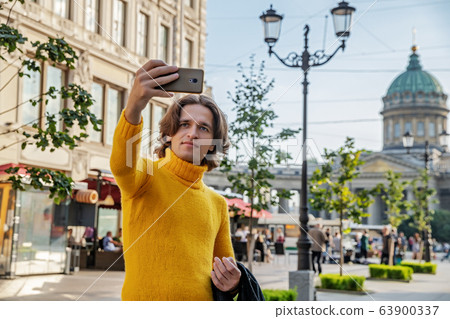 The handsome men does selfie, Look from outside, he dressed in a yellow sweater, a black raincoat or jacket is his hands, Bolshaya Konyushennaya street and Kazan Cathedral on background, sunny day The handsome men does selfie, Look from outside, he dressed in a yellow sweater, a black raincoat or jacket is his hands, Bolshaya Konyushennaya street and Kazan Cathedral on background, sunny day 63900337