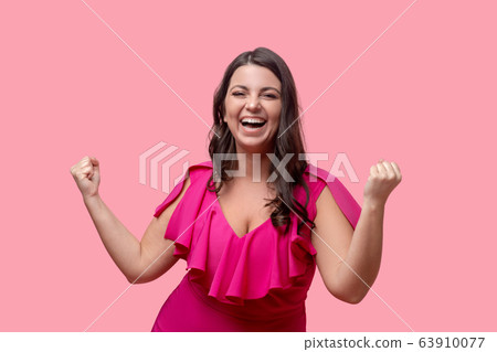 Dark-haired woman in a pink dress rejoicing clenching fists. 63910077