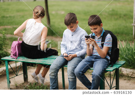 Two boys and girl use their phones during school breack. Cute boys sitting on the bench and play online games 63916130