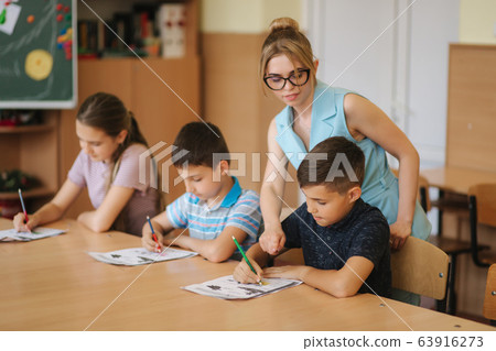 Teacher helping school kids writing test in classroom. education, elementary school, learning and people concept Teacher helping school kids writing test in classroom. education, elementary school, learning and people concept 63916273